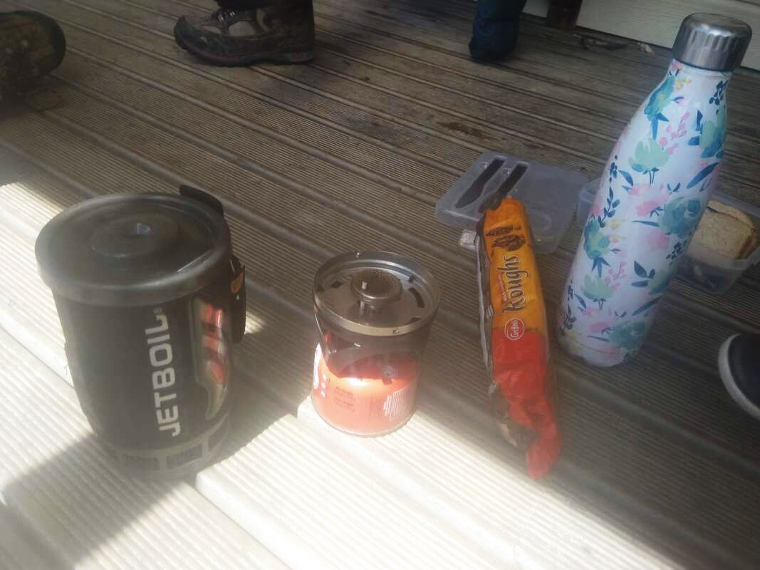 Picture of a picnic lunch on a wooden deck. There is a Jet Boil stove, a packaet of biscuits and a aluminum water bottle