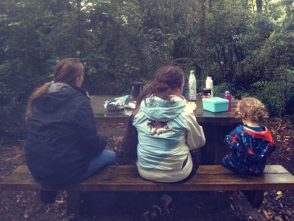 Picture of the backs of three children sitting at a picnic table. The children are wearing raincoats.