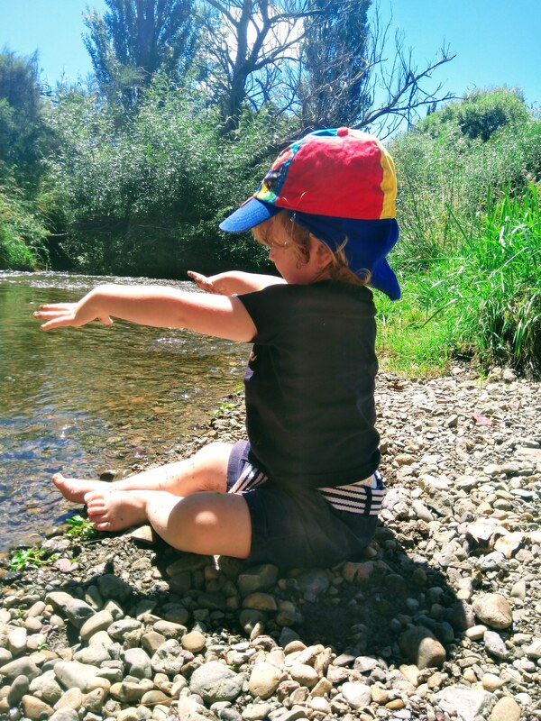 A toddler is sitting on the edge of a stream throwing a stone into the water.  The toddler is wearing a red hat and has no shoes on.