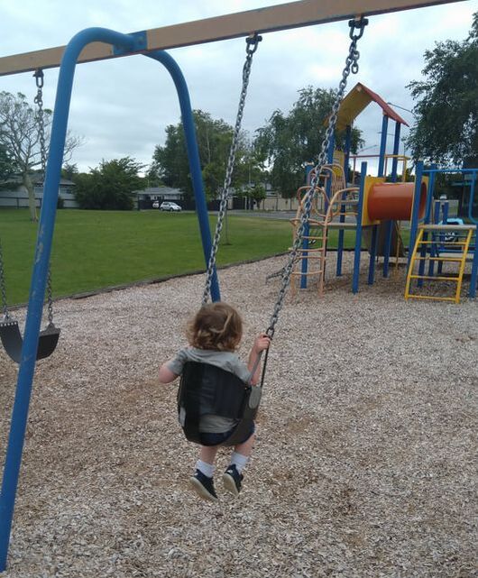 A small child swings on a swing set.  A play tower is in the background.  The child is wearing a green top and blue shoes. The swing is black and the frame is blue.
